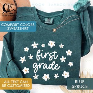 May include: Blue spruce Comfort Colors sweatshirt with white floral accents and the words "first grade." The sweatshirt has a crew neck and long sleeves. The text "ALL TEXT CAN BE CUSTOMIZED" is also visible.