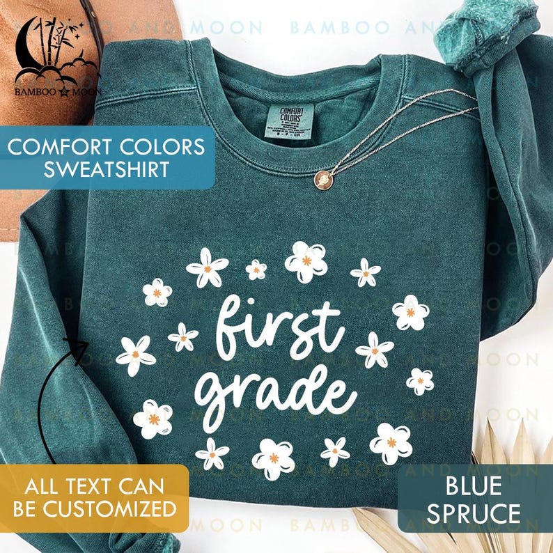 May include: Blue spruce Comfort Colors sweatshirt with white floral accents and the words "first grade." The sweatshirt has a crew neck and long sleeves. The text "ALL TEXT CAN BE CUSTOMIZED" is also visible.