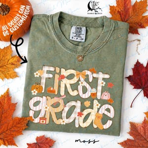 May include: An olive green t-shirt with a crew neckline and a graphic print that reads First Grade in large, light-colored plaid letters. The design includes small illustrations of pumpkins, flowers, leaves, a slice of pie, and a coffee cup.
