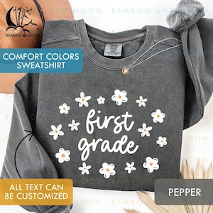 May include: A dark gray Comfort Colors sweatshirt with the words "first grade" in white, surrounded by white and orange flowers. The text "ALL TEXT CAN BE CUSTOMIZED" is also visible. The sweatshirt is pepper colored.