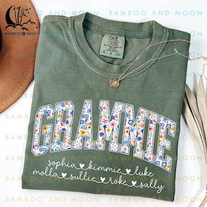 May include: A green t-shirt with the word "GRAMMIE" in white letters outlined in black. The letters are filled with a floral pattern in shades of pink, blue, yellow, and green. Below the word "GRAMMIE" are the names "sophia", "kimmie", "luke", "molla", "sullie", "roke", and "sally" written in white with hearts between each name.