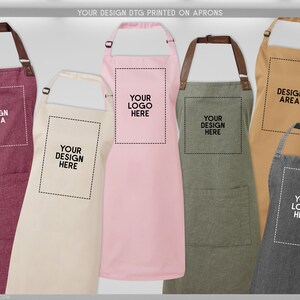 May include: A collection of aprons in various colors, including burgundy, cream, pink, olive green, tan, and charcoal. Each apron features a design area or logo placement, with text that reads "YOUR DESIGN DTG PRINTED ON APRONS".