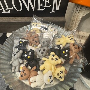 May include: A galvanized metal tray filled with small, individually packaged teddy bear figurines. The bears are in various colors, including black, brown, gray, and cream, with stitched details and cross-shaped eyes. A Halloween-themed sign is in the background.