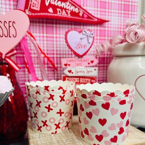 May include: Two decorative cupcake liners with scalloped edges. One liner features a pattern of red X's and pink O's on a cream background. The other liner has a pattern of red and pink hearts. Valentine's Day themed.