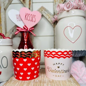 May include: Two Valentine's Day cupcake liners. One is red with white hearts and gold trim. The other is white with red wavy trim and gold text that reads "Happy Valentine's Day".