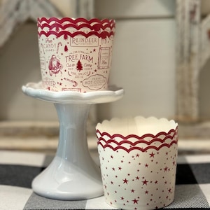 May include: Red and white cupcake liners with a Christmas tree farm design. The liners feature a red scalloped edge and a white background with red stars. The text on the liners reads "Reindeer Christmas Tree Farm Cut & Carry Cider Fine, Spruce and Fir Fresh Hot Cocoa".
