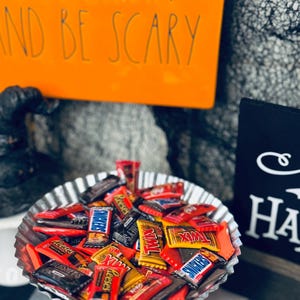 May include: A galvanized metal serving dish filled with assorted miniature candy bars, including Snickers, Twix, and Reese's. The dish sits on a gray pedestal. An orange sign with the text "EAT, DRINK AND BE SCARY" is in the background.