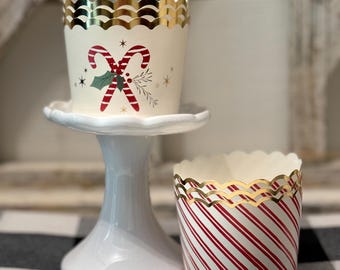 May include: Two decorative cupcake liners. One is cream-colored with a candy cane design and gold scalloped trim. The other is white with red diagonal stripes and gold scalloped trim. Both are festive and suitable for holiday baking.