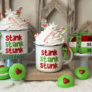 Puede incluir: Dos tazas blancas con las palabras "stink stank stunk" en rojo y verde, cubiertas con crema batida, bastones de caramelo y adornos temáticos de Grinch. También una caja de regalo y macarons verdes con detalles de corazón rojo.