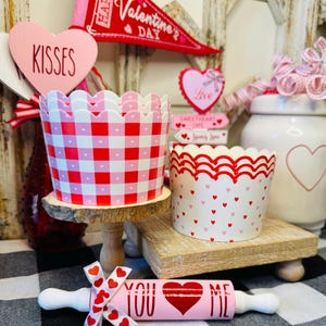 May include: Valentine's Day themed decorations including two paper cups, one with a red and white gingham pattern and the other with red hearts. A pink rolling pin with the words "YOU" and "ME" and a heart design is also present.