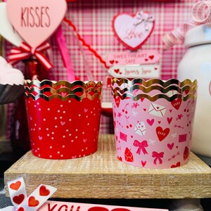 May include: Two Valentine's Day cupcake liners. One is red with white polka dots, the other is pink with hearts, bows, and envelopes. Both have gold scalloped edges. A pink rolling pin with "You & Me" text is in the foreground.