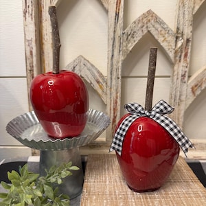 Pode incluir: Duas decorações de maçã vermelha brilhante com caules de madeira. Uma assenta num pedestal de metal galvanizado, a outra num bloco de madeira e adornada com um laço xadrez preto e branco. O fundo inclui uma moldura de janela branca desgastada.
