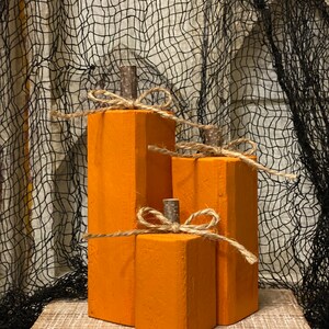 Peut inclure: Trois décorations de citrouilles en bois orange de tailles différentes. Chaque citrouille a une petite tige marron et un nœud en ficelle. Les citrouilles sont exposées sur une surface en bois devant un fond de filet noir.