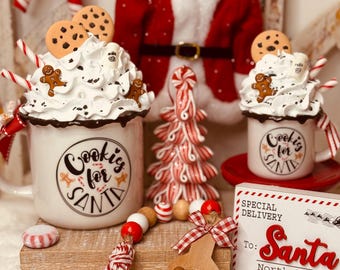 Puede incluir: Decoración navideña con tazas con crema batida, galletas y hombres de jengibre. Una cuchara de madera contiene más golosinas, y un letrero dice "Special Delivery To: Santa, North Pole". También se ve una guirnalda de cuentas rojas y blancas y un traje de Papá Noel.