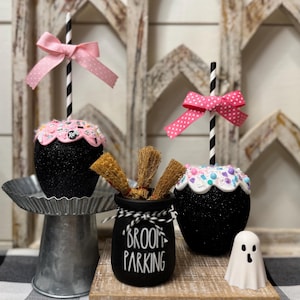 Può includere: Decorazioni a tema Halloween con due mele nere glitterate con fiocchi rosa e cannucce a righe. Sono presenti anche un piccolo vaso nero con la scritta "BROOM PARKING" e una statuina a forma di fantasma bianco. Le decorazioni sono su una superficie di legno.