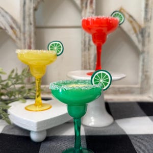 May include: Three colorful margarita glasses, in yellow, red, and green, are displayed. Each glass has a sugared rim and a lime wedge garnish. The glasses are on white pedestals and a checkered tablecloth.