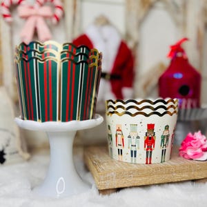 May include: Two decorative cupcake liners. One is green with red and white stripes, and the other is white with nutcracker illustrations. Both have gold scalloped edges. The liners are on a white pedestal and a wooden block, respectively. Christmas theme.