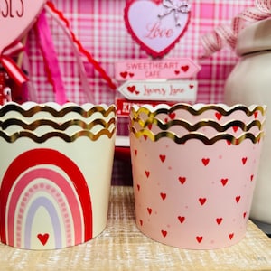May include: Two decorative cupcake liners. One is cream-colored with a red rainbow design and a gold scalloped edge. The other is pink with red hearts and a gold scalloped edge. Valentine's Day theme.