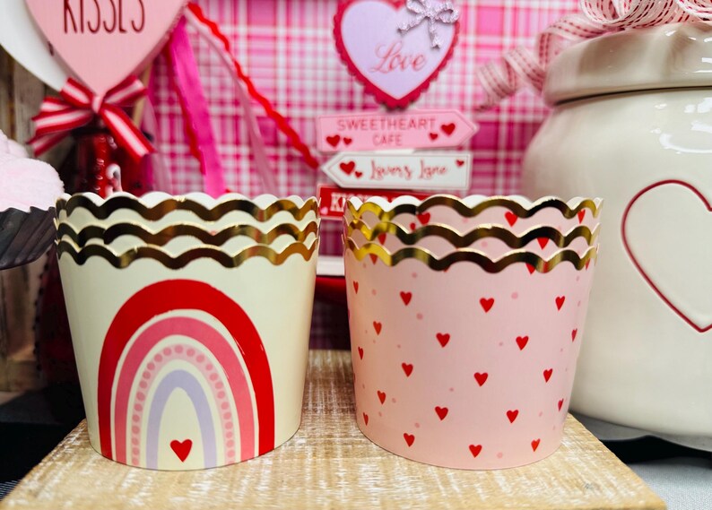 May include: Two decorative cupcake liners. One is cream-colored with a red rainbow design and a gold scalloped edge. The other is pink with red hearts and a gold scalloped edge. Valentine's Day theme.