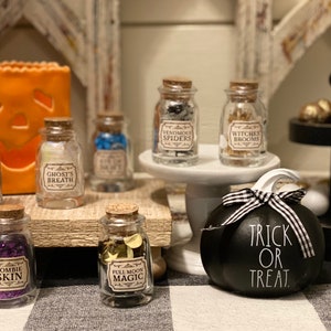Könnte beinhalten: Eine Halloween-Dekoration mit kleinen Glasbehältern mit Korken, beschriftet mit gruseligen Namen wie "Drachenschuppen" und "Zombieskin". Ein schwarzer Kürbis mit dem Text "Trick or Treat" und eine orangefarbene Jack-o'-Laterne sind ebenfalls vorhanden.