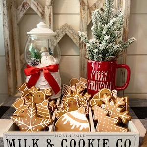 May include: A festive Christmas display featuring gingerbread cookies in a wooden tray with the text "Milk & Cookie Co.". A red mug with "Merry Christmas" and a snow globe with a snowman are also present.