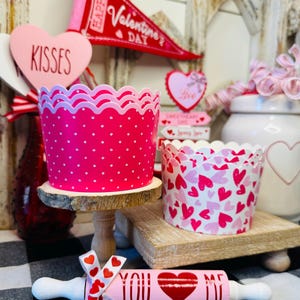 May include: Valentine's Day themed decorations. Includes pink polka dot and heart-patterned cupcake liners, a rolling pin with "YOU <3 ME" text, and a banner that says "Happy Valentine's Day".