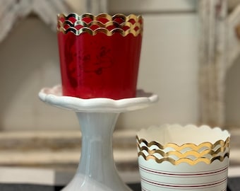 May include: Two red and white cupcake liners with gold scalloped edges. The liners are on a white pedestal cake stand. The stand is on a black and white checkered tablecloth.