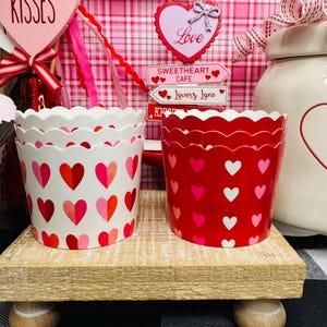 May include: Two decorative paper cups with scalloped edges sit on a wooden stand. One cup is white with red and pink hearts, the other is red with white and pink hearts. The background includes Valentine's Day decorations.
