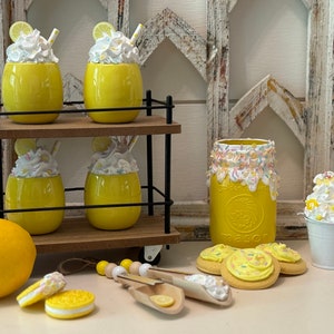 May include: A set of four yellow ceramic mugs with white whipped cream and lemon slices on top, displayed on a black metal trolley with wooden shelves. A yellow mason jar with white frosting and sprinkles is next to a white metal bucket with whipped cream and a lemon slice. Two yellow cookies with white frosting and sprinkles are on the table.