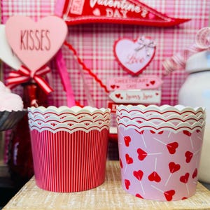 May include: Two Valentine's Day cupcake liners. One is red with white vertical stripes, the other is pink with red heart-shaped lollipop designs. Both have a scalloped white trim. The background includes Valentine's Day decorations.