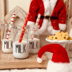 Pode incluir: Uma cena festiva de Natal com pequenas garrafas de leite de vidro com palhinhas listradas em vermelho e branco. As garrafas estão rotuladas com "MILK". Biscoitos com pepitas de chocolate em um pedestal branco e um gorro de Papai Noel vermelho em primeiro plano.