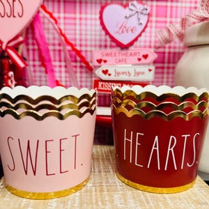 May include: Two decorative paper cups with scalloped edges and gold trim. One is light pink with the word "SWEET" in red, and the other is red with "HEARTS" in white. Valentine's Day themed.