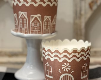 May include: Two brown cupcake wrappers with white gingerbread house designs. The wrappers have scalloped edges and are placed on a white pedestal and a black and white checkered surface. The wrappers are designed to look like gingerbread houses.