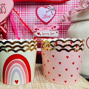 May include: Two decorative cupcake liners with gold scalloped edges. One is cream-colored with a red rainbow design and a heart. The other is pink with red hearts. Valentine's Day theme.