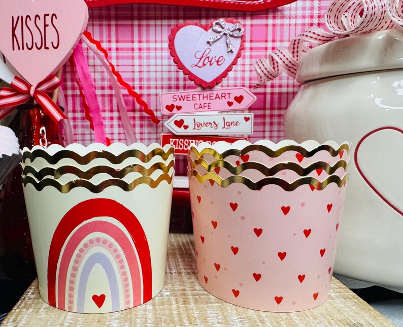 May include: Two decorative cupcake liners with gold scalloped edges. One is cream-colored with a red rainbow design and a heart. The other is pink with red hearts. Valentine's Day theme.