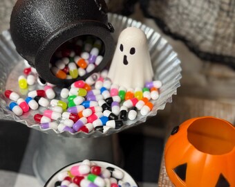 May include: A Halloween-themed still life featuring a black cauldron spilling colorful capsule-shaped candies onto a silver tray. A small white ghost figure and an orange jack-o'-lantern are also present. A green and white mug is filled with more candies.