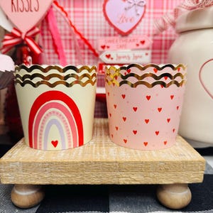 May include: Two decorative cupcake liners. One is cream-colored with a rainbow design, the other is pink with red hearts. Both have gold scalloped edges. They sit on a small wooden stand, with Valentine's Day decorations in the background.