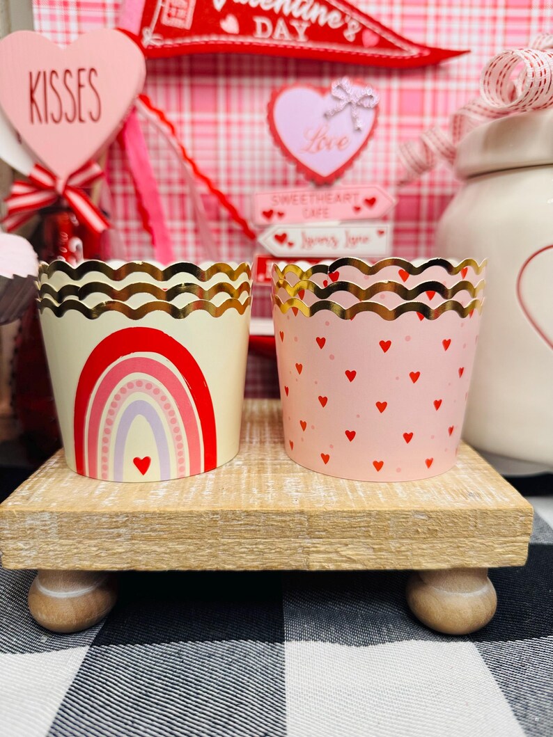 May include: Two decorative cupcake liners. One is cream-colored with a rainbow design, the other is pink with red hearts. Both have gold scalloped edges. They sit on a small wooden stand, with Valentine's Day decorations in the background.
