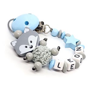 May include: A baby pacifier clip with a light blue and gray color scheme. The clip features a gray wolf-shaped charm, a crocheted ball, and a star-shaped bead. The name "LEO" is spelled out with white letter beads.