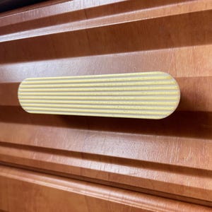 May include: A gold and cream-coloured cabinet pull with a grooved design. The rectangular handle is mounted on a wooden cabinet with horizontal grooves. The pull is a decorative hardware element.