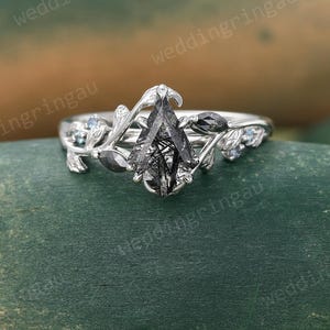 May include: A pear-shaped black diamond engagement ring with a delicate floral design. The ring features a silver band with two small black diamonds and two small blue diamonds on each side of the main stone.