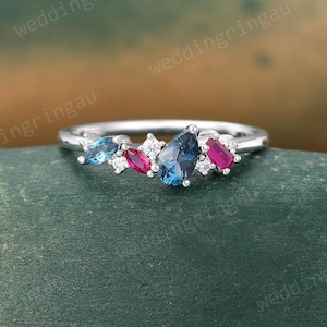May include: A silver ring with a cluster of gemstones in shades of blue, red, and clear. The ring features a central dark blue stone, with smaller blue and red stones on either side. The ring is set against a green and brown background.