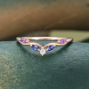 May include: A rose gold wedding band with a V-shaped design featuring a small diamond and three marquise-shaped gemstones: two purple and one blue.