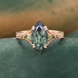 May include: A rose gold ring with a large, teal-green marquise-cut gemstone at its centre. Two smaller, pear-shaped, green gemstones are either side of the main stone. The band has an ornate, filigree design. The ring is set against a blurred, green and brown background.