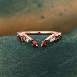 Puede incluir: Un delicado anillo de oro rosa con un diseño en forma de V adornado con piedras preciosas rojas y blancas. El anillo está sobre un fondo verde.