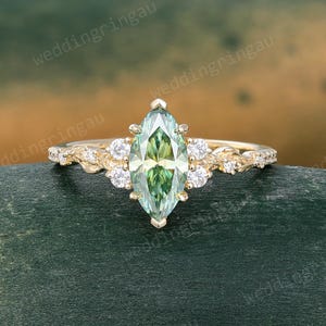 May include: A gold ring featuring a central marquise-cut green gemstone held by prongs. The band has a textured, branch-like design adorned with small round clear stones on both sides of the centre stone. The text weddingringau is visible.