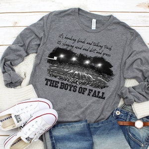 May include: A grey long-sleeved t-shirt with a black and white image of a football stadium and the text "It's knocking heads and talking trash. It's slinging mud and dirt and grass. The Boys of Fall."