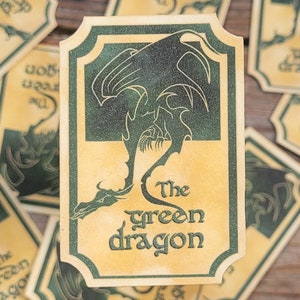 Peut inclure: Gros plan sur plusieurs petits autocollants rectangulaires de couleur crème avec un motif de dragon vert. Les autocollants comportent le texte "The Green Dragon" dans une police de caractères stylisée.