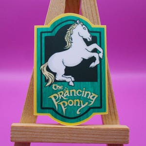 May include: A rectangular sign with a green border and a black background featuring a white horse rearing up. The text "The Prancing Pony" is in a teal font at the bottom. The sign is displayed on a small wooden easel.