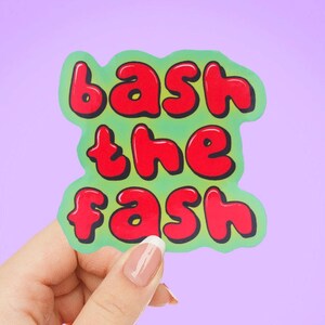 May include: A green sticker with the words "bash the fash" in red bubble letters. The sticker is held by a hand against a purple background. The text has a white outline.